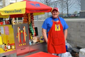 Man in blue shirt with red apron standing by a Vienna Beef umbrella and a hot dog stand, he doesn't look like he is losing his business